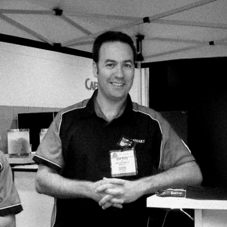 Black-and-white candid of Adam Rozen at a trade-show booth, wearing a branded polo and exhibitor badge